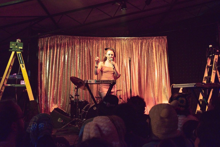 Mid shot from the crowd of Neggy Gemmy performing on stage holding the mic and raising her left hand.