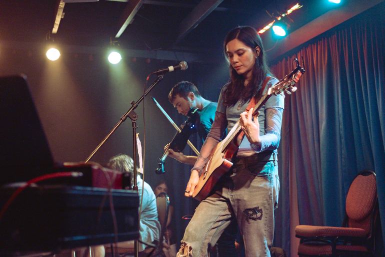 Maya, of Chanel Beads, stands off center and to the right of the frame in focus. She has an electric guitar hanging with a strap around her neck and is looking down at it while playing. To the left