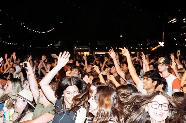 The image is of a large crowd, most having their hands up and dancing. At the front, you can see several women smiling, one in a hat, one in a black shirt, and another in glasses. At the top of the image you can see string lights across the venue.