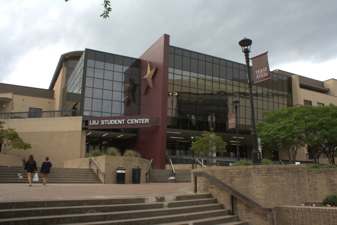 Exterior view of the LBJ Student Center at Texas State University with glass windows, a large star emblem, and two people walking up the steps.