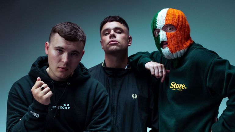 The members of the Irish rap group Kneecap pose together; the one on the right being DJ Próvaí wears a green, white, and orange balaclava, symbolising the Irish flag.