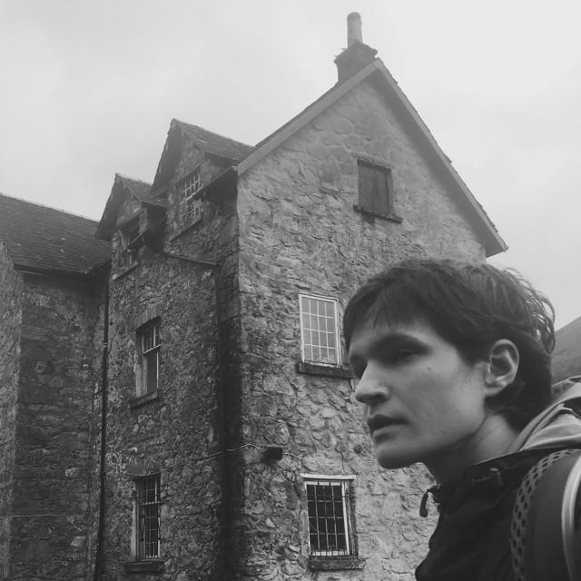 The image is a black and white photo of part of a large stone house, in the bottom left corner, a woman in a hoodie with short hair is looking away from the camera. 