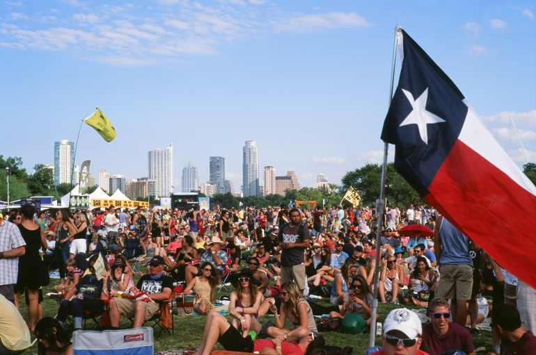 A large crowd gathered at the Austin City Limits music festival in front of the main stage, with the skyline of downtown Austin, Texas, visible in the background at sunset. A large balloon in the left hand corner floats above the crowd with “Austin City Limits” titled on it. Description: The photo captures a vibrant moment from the Austin City Limits (ACL) Music Festival, held in Zilker Park, Austin, Texas. Thousands of people face a massive concert stage backed by a glowing skyline of modern high-rise buildings during the day's golden hour. A prominent balloon reading “Austin City Limits” hovers above the crowd.