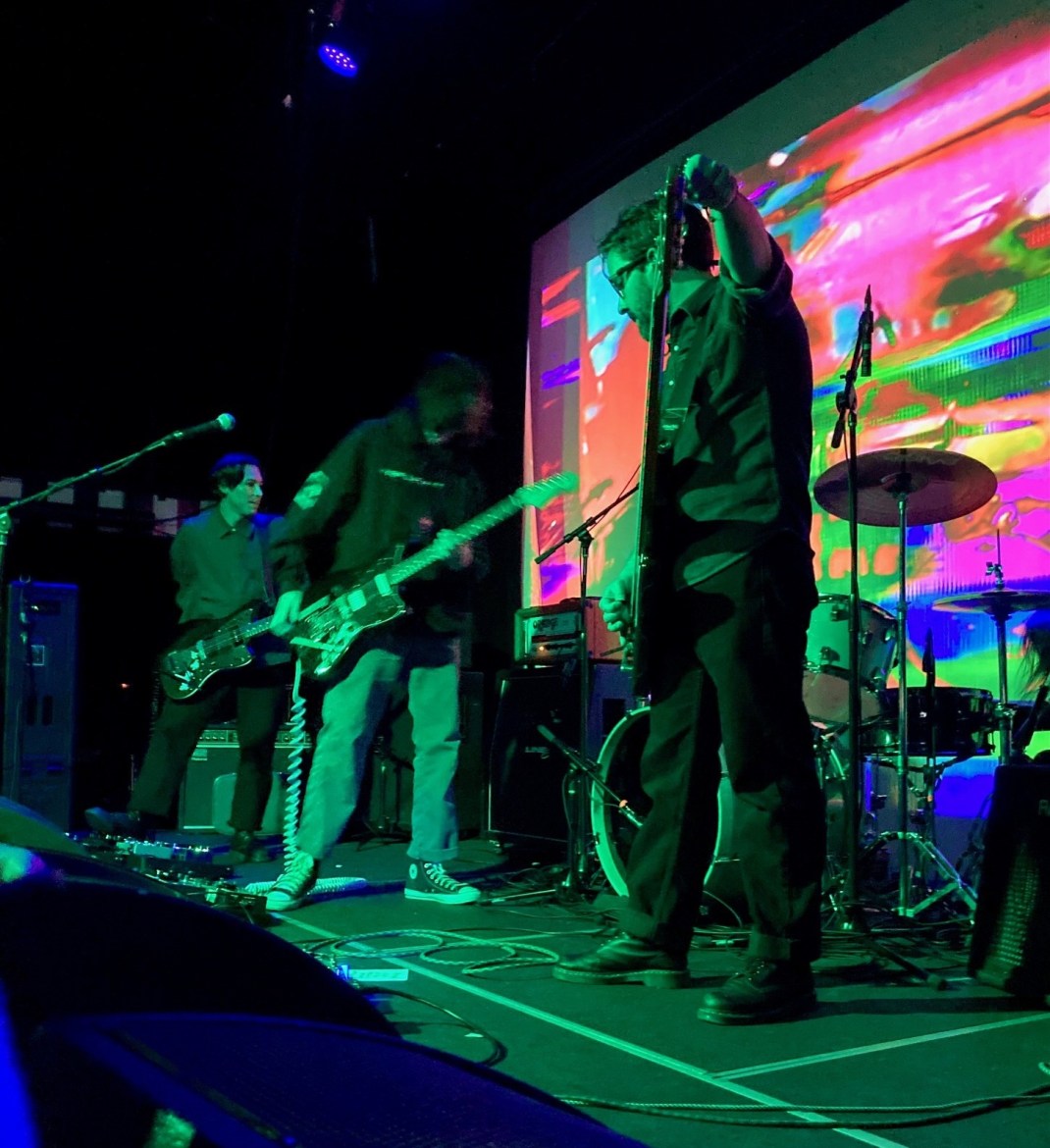 All members of the band pictured in green lighting, playing live music. There is one man with glasses, his left hand holding the microphone. To his left is the guitarist, his face is blurry due to the camera quality. The bassist is semi-covered by the guitarist. They are all looking away from the camera. Behind them is a drum set, though the drummer is obscured.