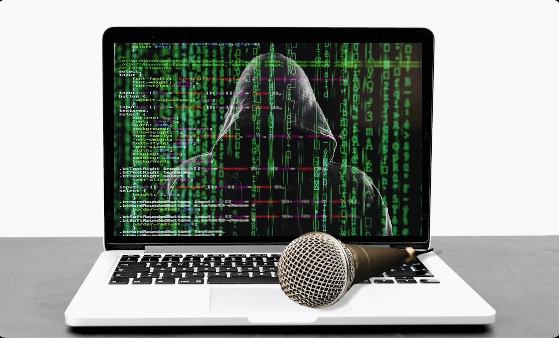 There is an open laptop sitting in the middle of a desk with a white wall in the background. On the screen, there is an image of code with binary numbers and a hooded figure in the background. There is a black microphone sitting on the keyboard portion of the laptop