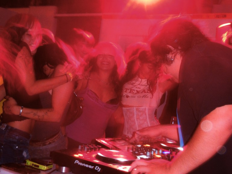 A DJ performs on Pioneer turntables while a crowd of people dance and embrace under red lighting.