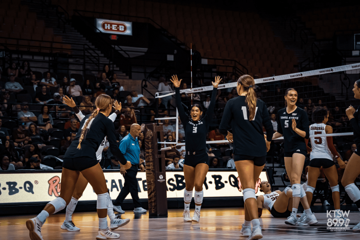 Texas State volleyball players celebrate a point at the net, smiling and raising their arms, while an opposing player sits on the court.
