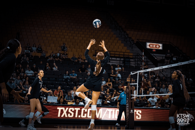 Volleyball player jumps to set the ball during a Texas State match, with teammates ready at the net and fans in the background.