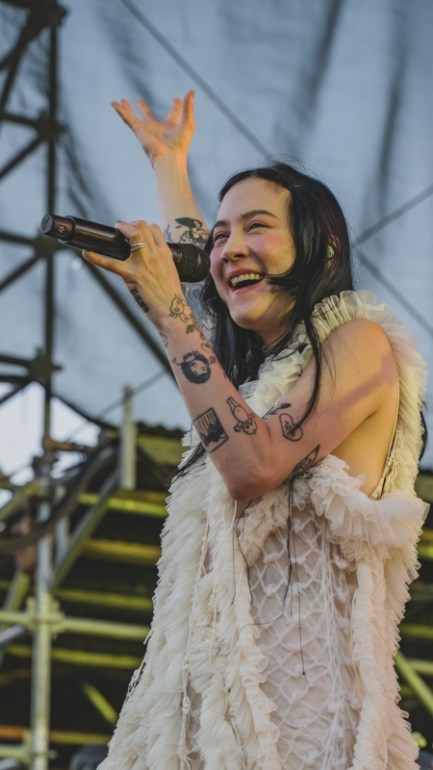 Michelle Zauner of Japanese Breakfast smiles while singing into a microphone on stage, with her hand raised in the air.