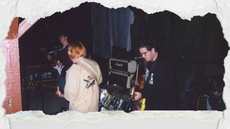 P.E.E. performing a concert in front of a crowd in an industrial setting. The picture has been edited to look ripped from a paper. 