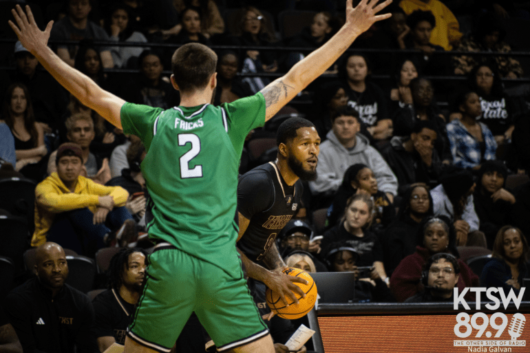 Texas State men’s basketball player holds the ball while a Marshall defender contests with arms raised in front of a crowd.