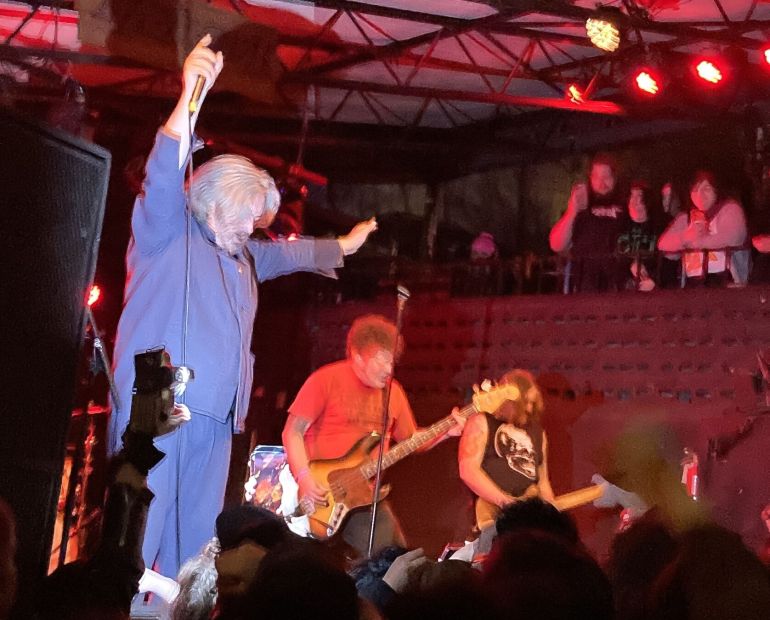 The photo shows a medium side shot of the vocalist, guitarist, and bassist of Orchid onstage while red lights shine over them. A flash shines over the vocalist Theo Mindell as he holds a microphone in his right hand, and the heads of people in the crowd can be seen on the bottom of the photo.