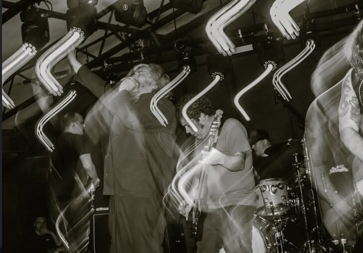 The photo shows a black and white shot of Orchid’s vocalist screaming into the microphone while their bassist and drummer are seen playing in the background. The lighting shows a shutter drag sort of effect, creating blurs of light around the band.