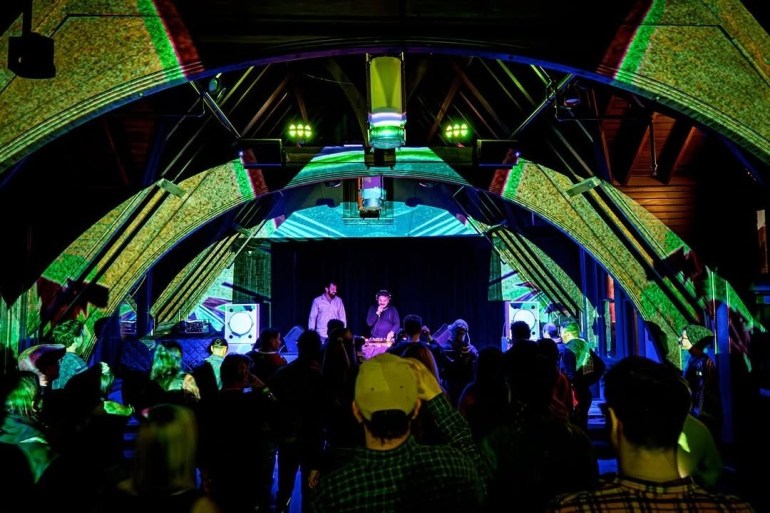 Inside the middle of a set, lasers are pointing at the ceiling with a medium-sized crowd in attendance.
