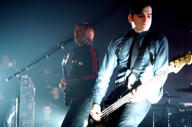 The band Interpol is playing a show. Bassist Carlos Dengler is featured on the right looking down at his bass while playing. Lead singer Paul Banks is on the left currently singing and looking at the crowd. The drums are in the bottom right corner, the audience in the bottom left.
