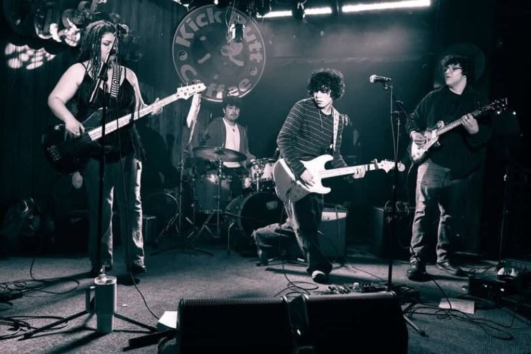 The photo shows a black and white image of the four members of metalloid playing on a stage with two microphones and monitors set up in the front and two amps set up on the back by the drum kit. Behind the drummer is a circular sign with the words, “Kickbutt Coffee."