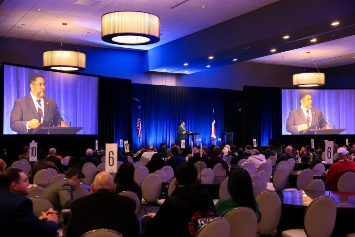 Texas AFL-CIO President Leonard Aguilar speaks at the 2026 COPE Convention. He is projected on two screens, as well as standing on stage, speaking in front of a large audience.