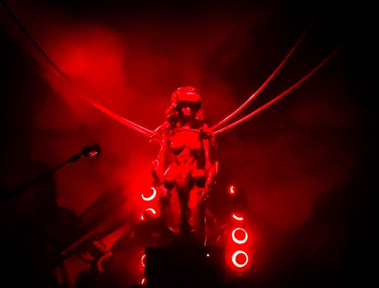 The image shows a statue of a robotic woman with cables coming out of her shoulders and a red light shines on it