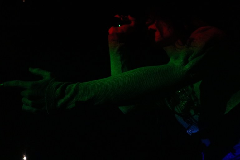 Performer sings into a microphone while reaching toward the crowd under red and green stage lighting.