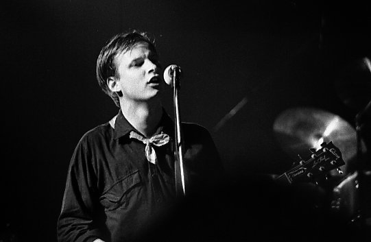 Black and white photograph of Andy Partridge singing on stage alone 