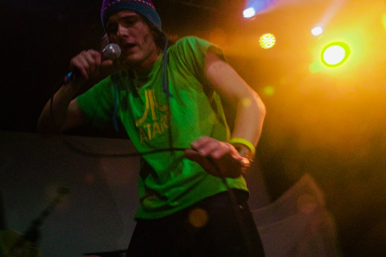 Performer in green shirt and beanie leans forward singing into a microphone under colorful stage lights.