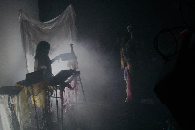 Electronic musician performs behind keyboard setup in heavy fog with bright backlighting and minimal visibility.