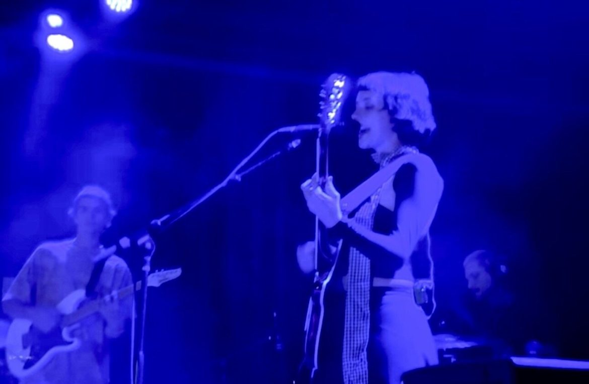 The band winter playing a show on stage. The stage is illuminated in blue light. Samira winter the lead singer/guitarist is center stage singing into the microphone. Another guitarist is seen to the left of her, and the drummer is playing the drums behind to the right of the singer. Image Credit: Paxton Poole (from when I saw her live)