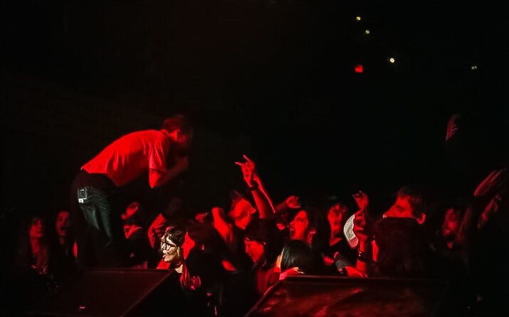 The image features a side shot of vocalist Jordan Dreyer screaming into the mic at the crowd below on the right side. The bottom of the image has monitors peaking out and there is red lighting shining on everyone.
