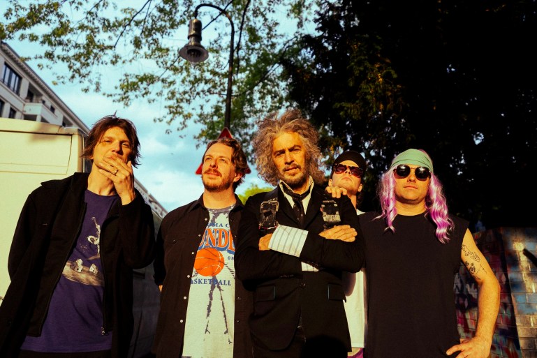 Five members of The Flaming Lips stand outdoors at sunset, facing the camera in warm golden light, with trees and buildings in the background.