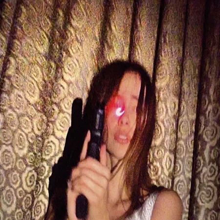 A stylized, low-light portrait of a woman standing in front of patterned curtains. She holds what appears to be a prop gun while a bright red light effect is visible near one eye, creating a dramatic, cinematic aesthetic with noticeable image grain.