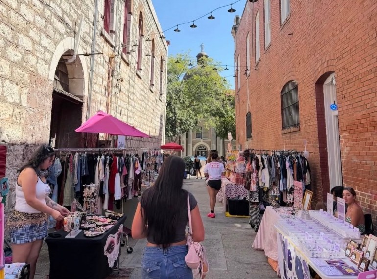 Group gathered in an alleyway that is filled with booths and speakers that display varying trinkets and handcrafted items.