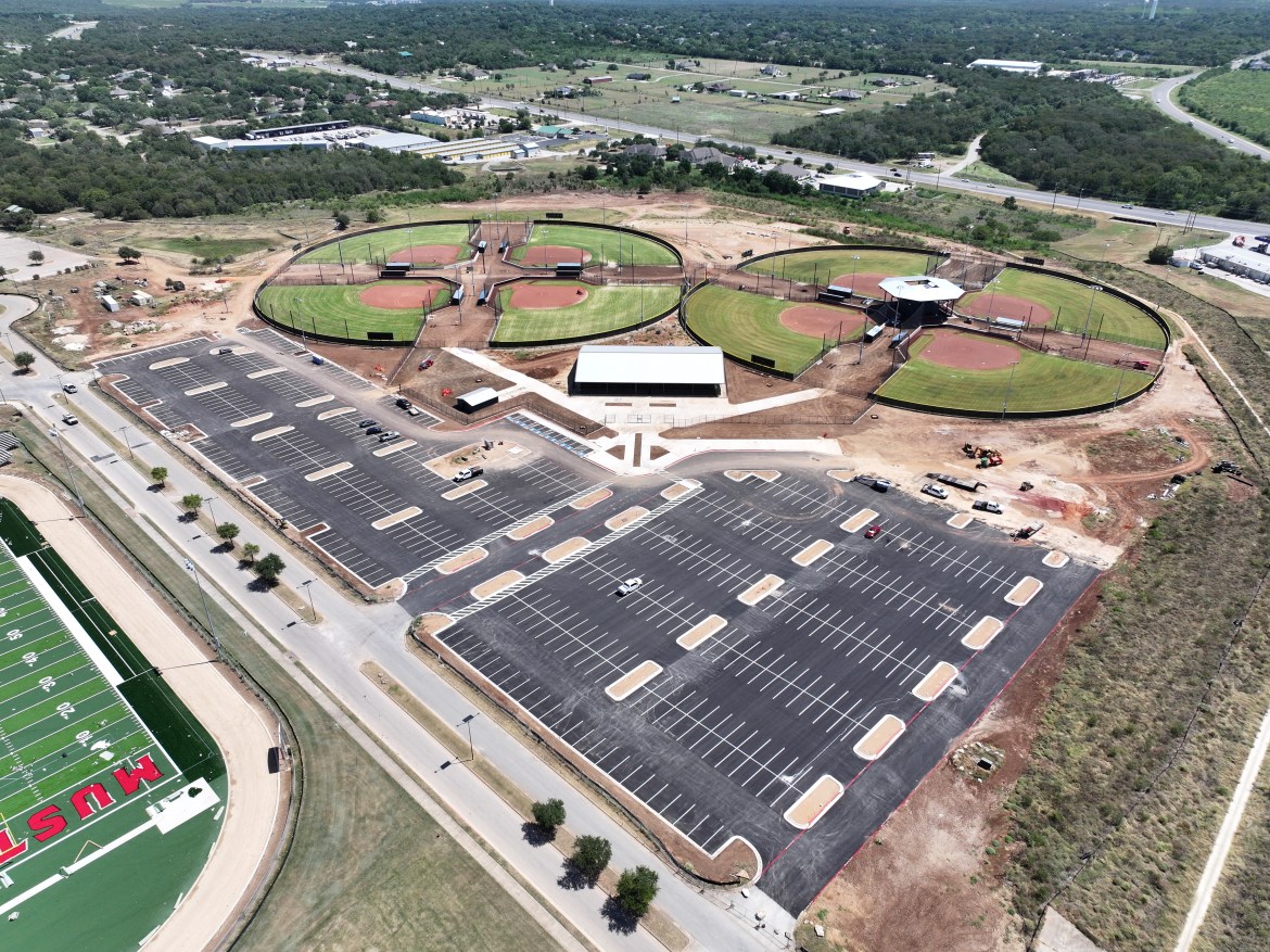 Aerial picture of Buda's Championship Park, featuring two sports fields and a parking lot. To the sides there is empty land with sand where the venue will be expanded to. At the bottom left there is an excavator working the land.