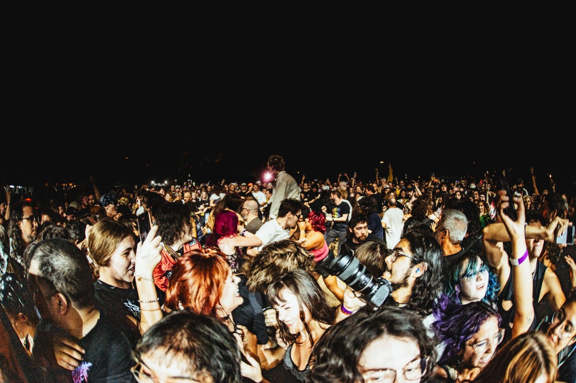 The image features a wide lens shot of people in the crowd moshing at night while a bright white light illuminates them.