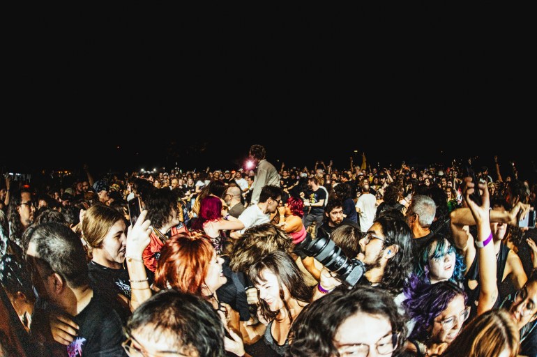 The image features a wide lens shot of people in the crowd moshing at night while a bright white light illuminates them.