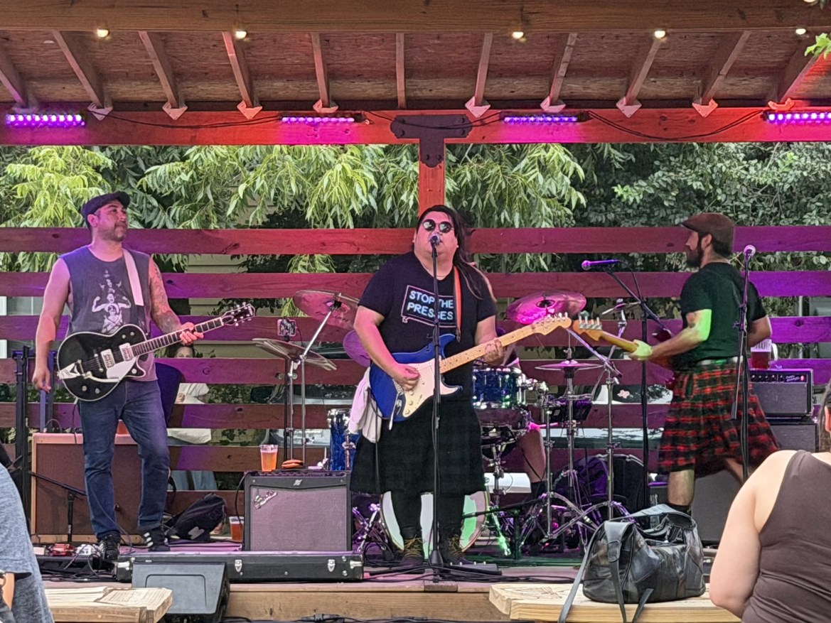 Three performers on a stage. The left musician is playing guitar, the middle musician is playing guitar and singing, the right musician is playing bass guitar.