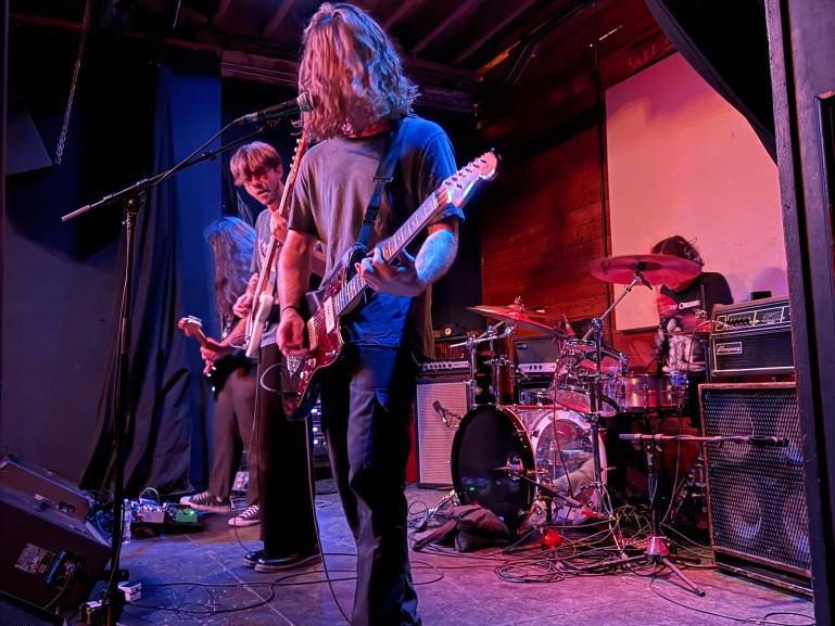 Four piece band playing on stage. The lead singer and guitarist looked at the camera with hair covering his face. Wide shot of the indoor stage at Mohawk