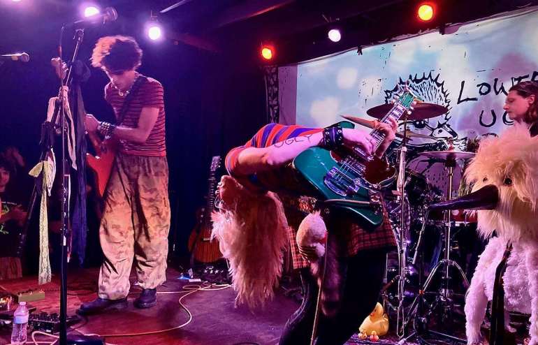 The image features Olivia Osby in the center of the stage bending over backwards playing a green guitar while Avsha Weinberg stands further back playing a red guitar. There are two microphones towards the left of the image and two pedal boards and cables scattered across the floor