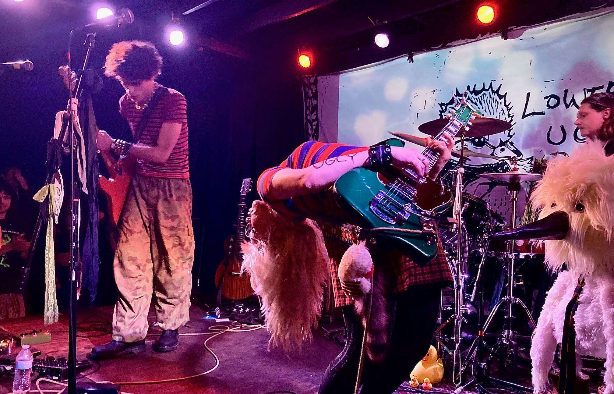 The image features Olivia Osby in the center of the stage bending over backwards playing a green guitar while Avsha Weinberg stands further back playing a red guitar. There are two microphones towards the left of the image and two pedal boards and cables scattered across the floor