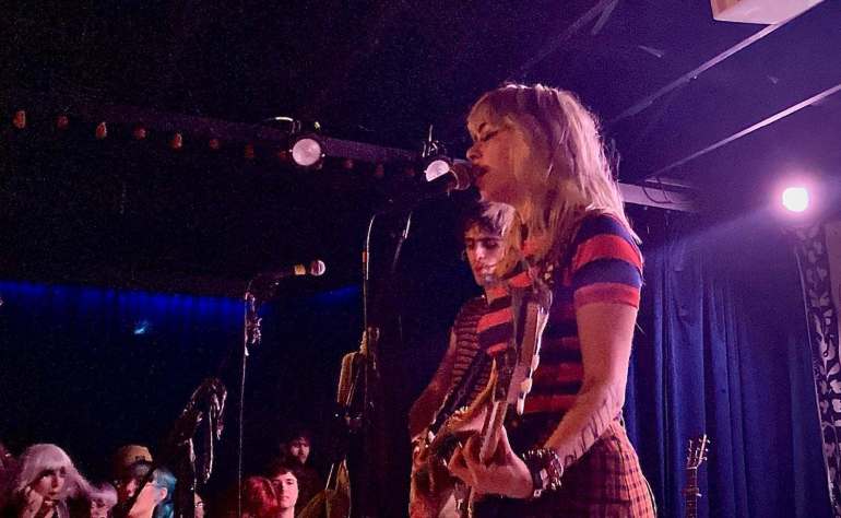  the image features a medium side shot of Olivia Osby singing into a mic while playing a guitar while the room is dim and lights from the stage shine on her.