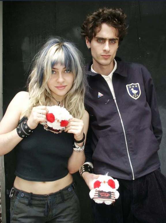 The image features Olivia Osby standing on the left in a black tank top and Avsha Weinberg on the right in a black jacket, both posing with a small white duck plushie with red cheeks and a striped shirt that says “Lowertown”. 