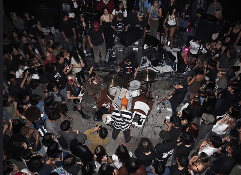 The image shows an overhead shot of the full band facing each other with a crowd surrounding them and a flash from the camera shines above them.