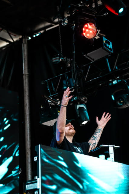 DJ 'Cozy Kev' is seen lifting his hands up while he is playing a set at the San Marcos Block Party. He stands in front of an LED screen that displays blue graphics.