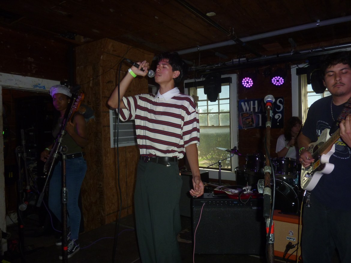 A boy, Deyna Reyna, sings into a microphone with his eyes closed while in front of his small band inside of a venue’s stage. There are lights behind him where his drummer sits.