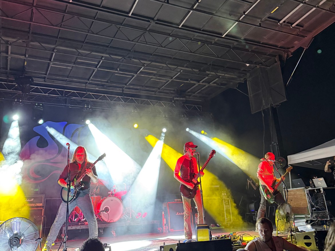 Rock band with 3 front facing members playing guitar and singing on stage underneath yellow and white lights at night. 