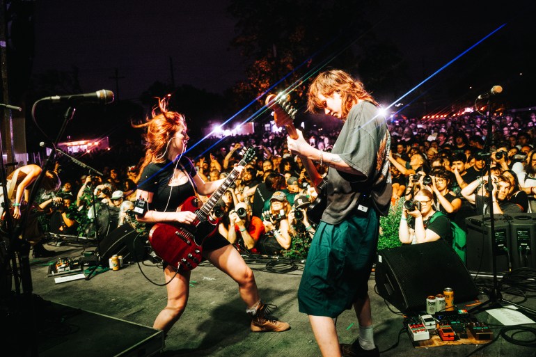 On stage is Ellie and Ava from Die Spitz both playing guitar facing each other with a crowd of people watching.