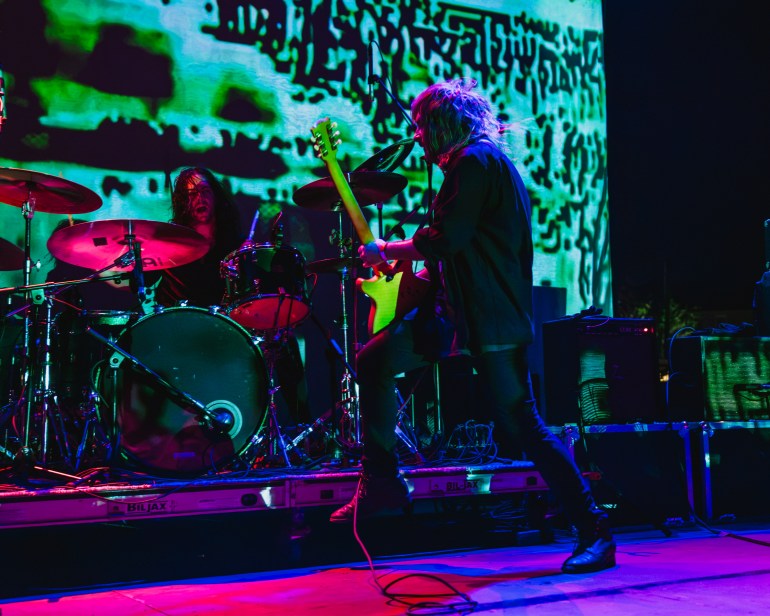 The front man of Uncle Acid and The Deadbeats is shown playing with his back turned facing the drummer under dark colored lights.