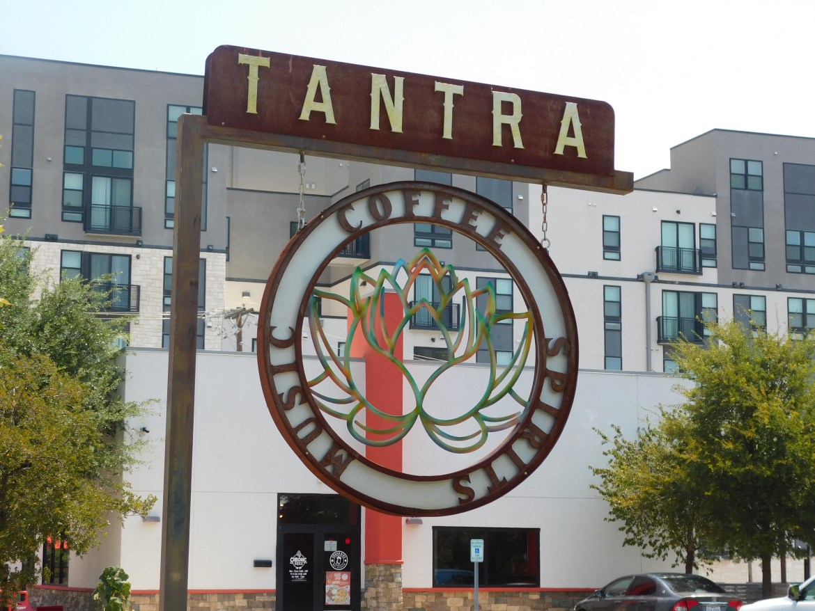 TANTRA written on a signpost with a logo of a lotus encapsulated by a circle that reads, “COFFEE SPIRITS MUSIC.”
