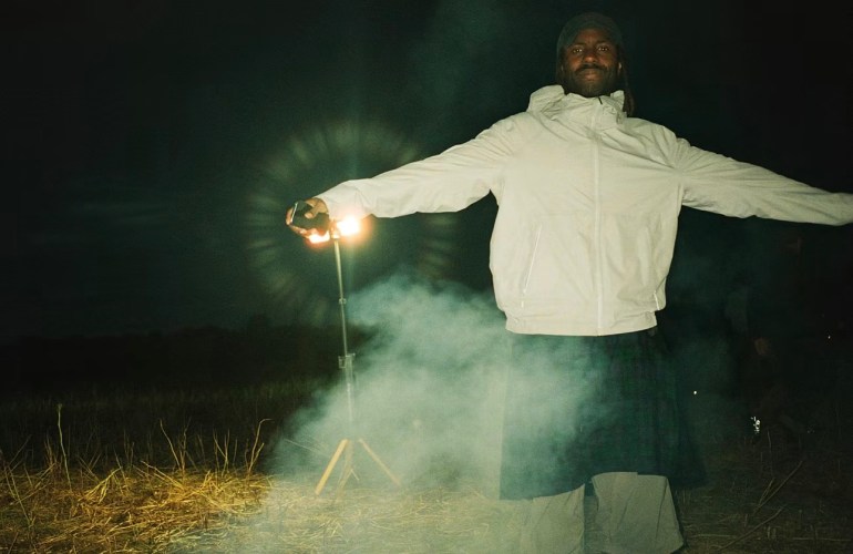 The image depicts Dev Hynes, who is the leading member of the band Blood Orange standing in a grass field with his arms out and what appears to be a light in his hand. He is holding out the light with smoking emitting around him.