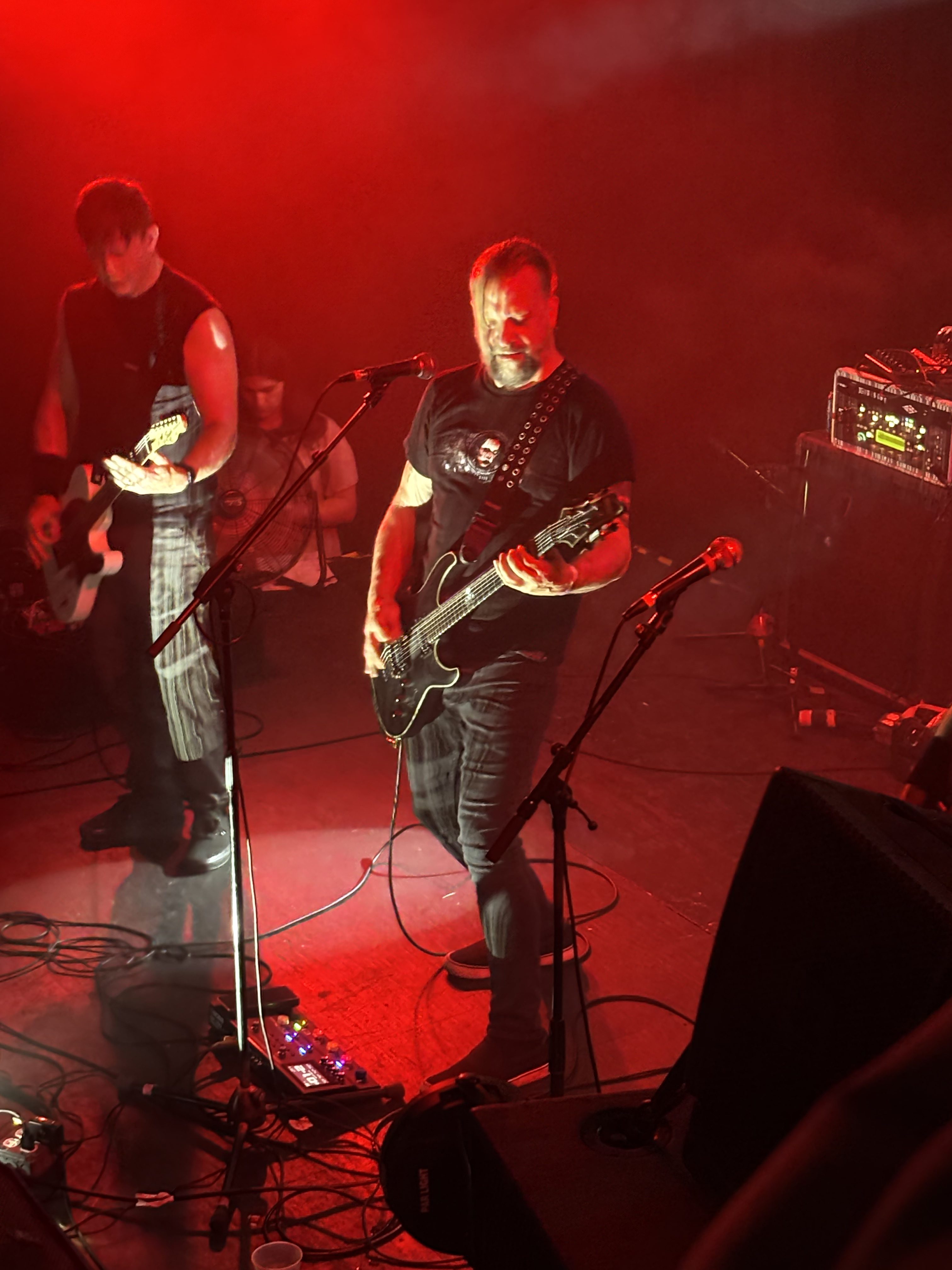 Acumen Nation supporting guitarist on the left of a stage, then frontman and guitarist Jason Novak beside a microphone and playing guitar on the right side of a red-lit stage 