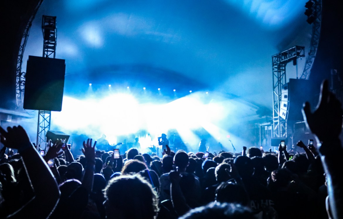 The image shows a wide shot of the crowd in the foreground shrouded in a bright blue light and the band is shown in the background with large speakers hanging on the sides of the stage.